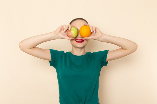 Front view young female in dark green shirt and blue jeans holding apple and orange on beige