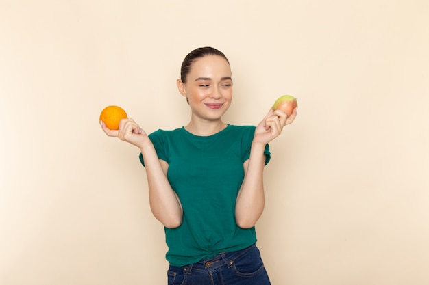 Front view young female in dark green shirt and blue jeans holding apple and orange smiling on beige