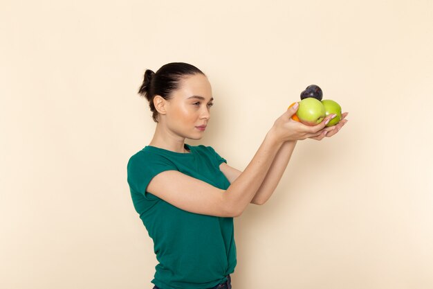 ダークグリーンのシャツとベージュにリンゴとオレンジを保持しているブルージーンズの正面の若い女性