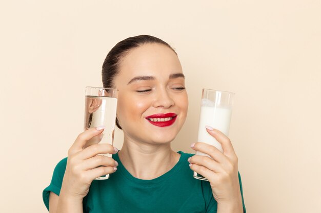 Front view young female in dark green shirt and blue jeans holding both milk and water smiling on beige