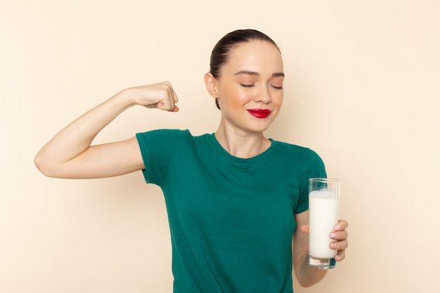 Front view young female in dark green shirt and blue jeans holding glass of milk flexing on beige
