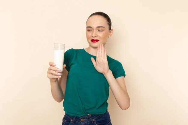 濃い緑色のシャツとベージュで飲むことを拒否する牛乳のガラスを保持しているブルージーンズの正面の若い女性