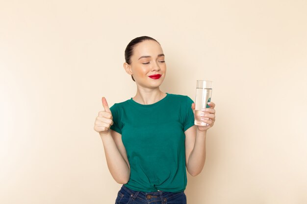 Front view young female in dark green shirt and blue jeans holding glass of water on beige