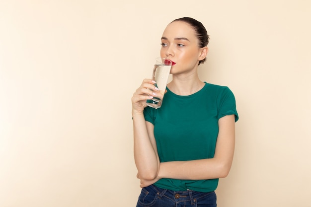濃い緑色のシャツとベージュで飲む水のガラスを保持しているブルージーンズの正面の若い女性