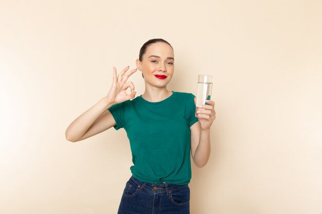 Front view young female in dark green shirt and blue jeans holding glass of water smiling on beige