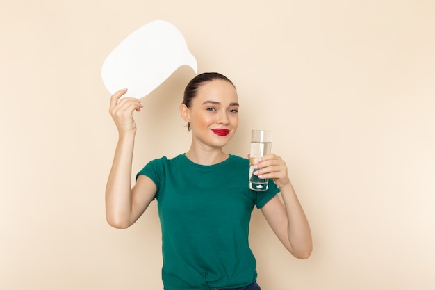 Front view young female in dark green shirt and blue jeans holding glass of water and white sign on beige