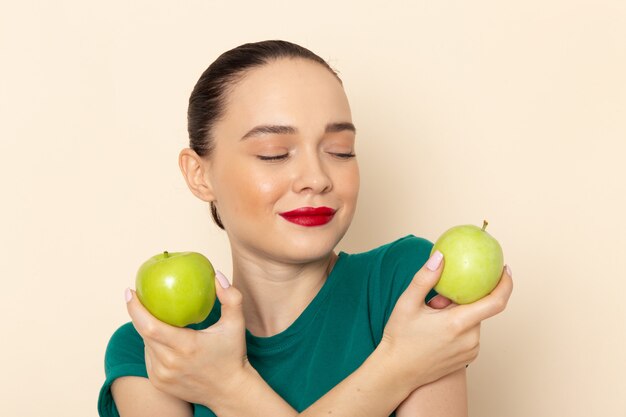 Front view young female in dark green shirt and blue jeans holding green apples with smile on beige