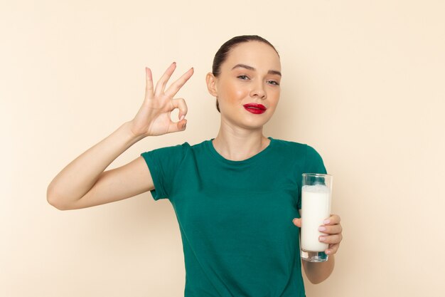 Front view young female in dark green shirt and blue jeans holding milk smiling on beige