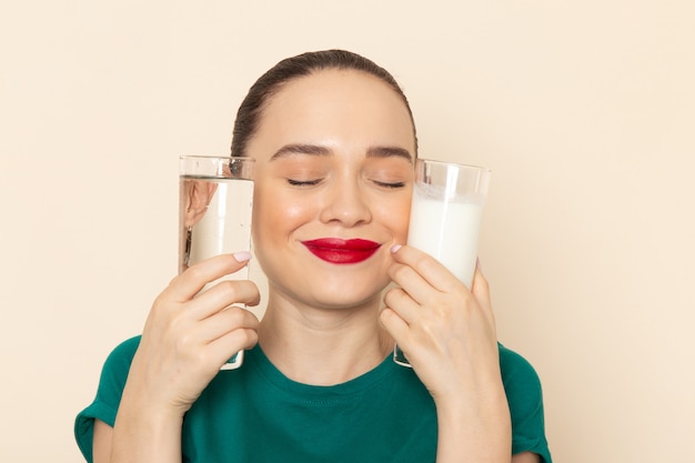 Front view young female in dark green shirt and blue jeans holding milk water smiling on beige