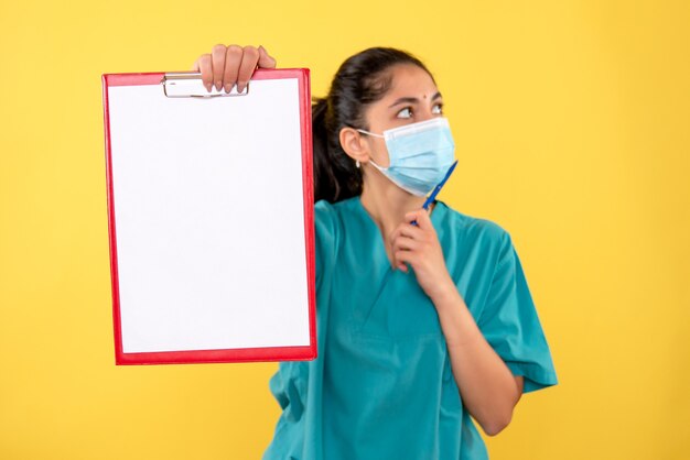 Front view of young female doctor holding clipboard on yellow wall