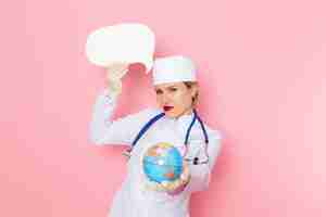 Бесплатное фото Вид спереди молодая женщина-врач в белом медицинском костюме с синим стетоскопом держит глобус и белый знак на розовом космическом медицинском госпитале