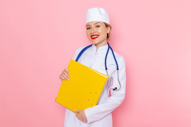 Бесплатное фото Вид спереди молодая женщина-врач в белом костюме с синим стетоскопом, держащая желтые файлы на розовом пространстве женской работы