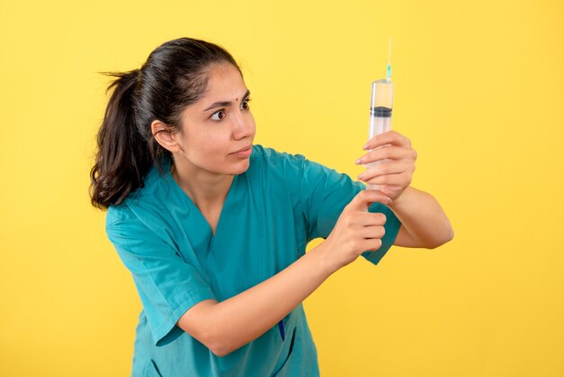 Front view of young female doctor looking at syringe on yellow wall