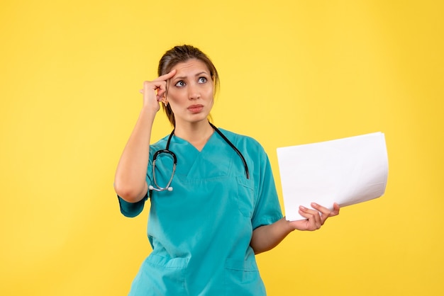 Вид спереди молодая женщина-врач в медицинской рубашке держит анализ бумаги и думает на желтом фоне