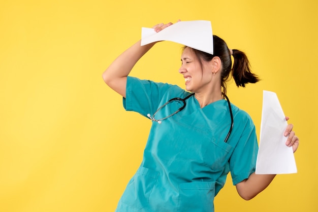 Giovane medico femminile di vista frontale in camicia medica che tiene analisi della carta su fondo giallo