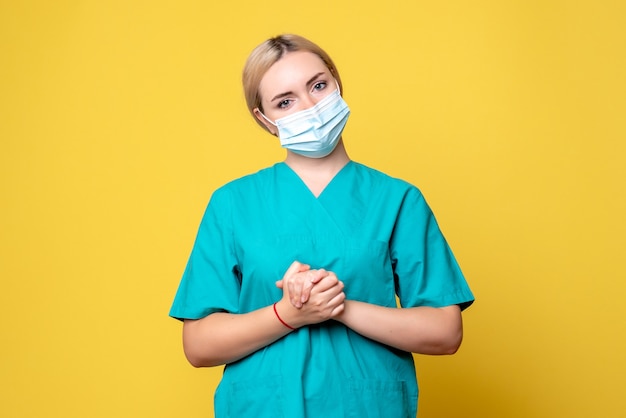 Vista frontale del giovane medico femminile in camicia medica e maschera sulla parete gialla