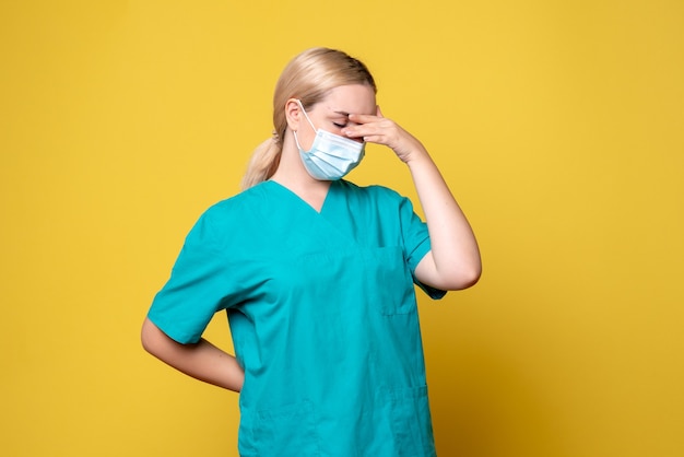 Front view of young female doctor in medical shirt and sterile mask feeling stressed on yellow wall