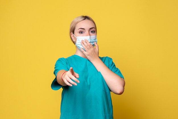 Front view of young female doctor in medical shirt and sterile mask on a yellow wall