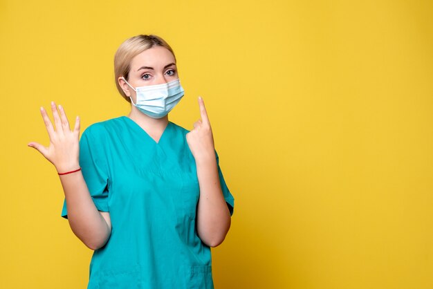 Front view of young female doctor in medical shirt and sterile mask on yellow wall