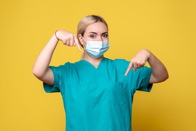 Front view of young female doctor in medical shirt and sterile mask on yellow wall