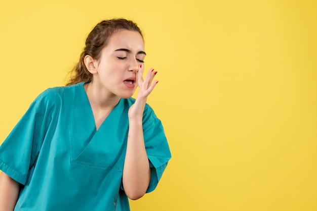 Front view of young female doctor in medical suit cleaning her nose on yellow wall