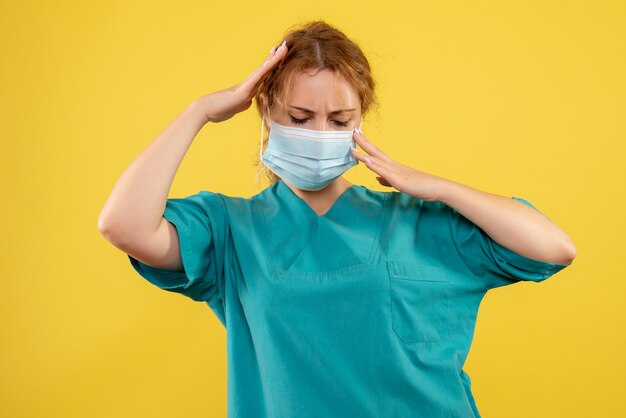 Front view of young female doctor in medical suit and mask having headache on yellow wall