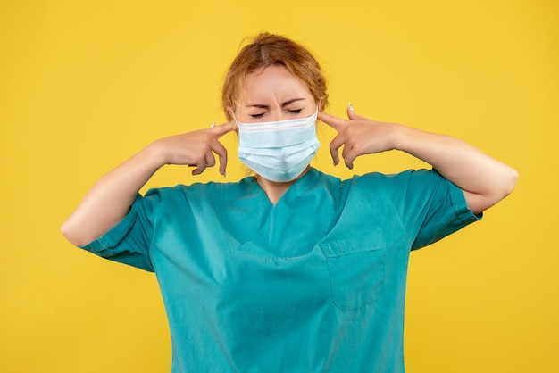 Front view of young female doctor in medical suit and mask on yellow wall