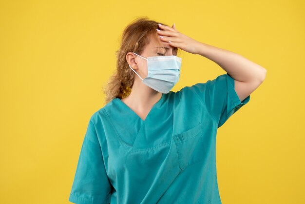 Front view of young female doctor in medical suit and mask on yellow wall