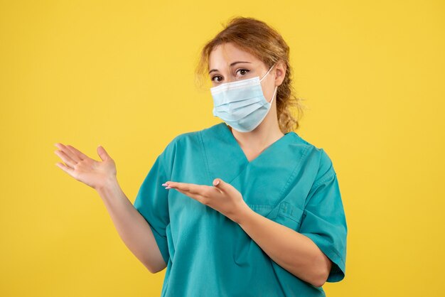 Вид спереди молодой женщины-врача в медицинском костюме и маске на желтой стене