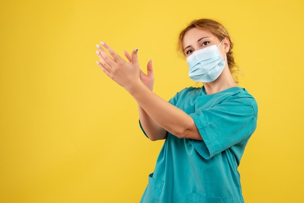 Front view of young female doctor in medical suit and sterile mask clapping on yellow wall