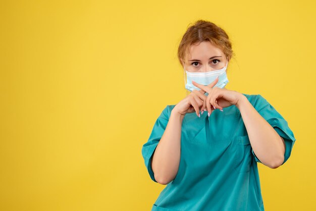 Front view of young female doctor in medical suit and sterile mask on a yellow wall