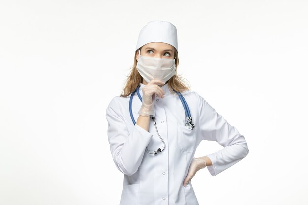 Front view young female doctor in medical suit wearing protective mask due to coronavirus thinking on white surface