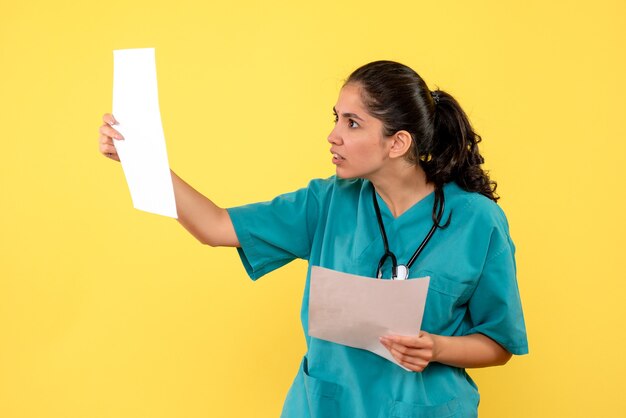 Front view of young female doctor in uniform checking documents on yellow wall