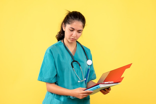 Front view of young female doctor in uniform looking at clipboard on yellow wall