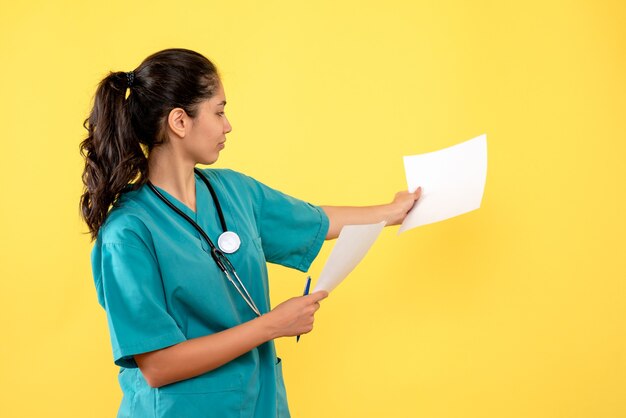 노란색 벽에 문서를보고 제복을 입은 젊은 여성 의사의 전면보기