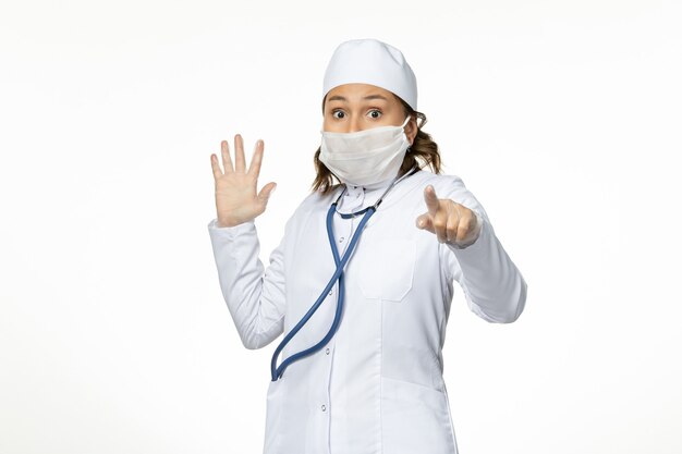 Front view young female doctor wearing protective sterile mask due to coronavirus scared on white surface