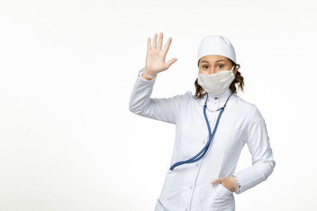 Front view young female doctor wearing protective sterile mask due to coronavirus showing her hand on white desk