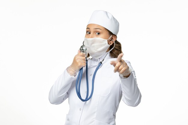 白い机の上で聴診器を使用してコロナウイルスによる保護滅菌マスクを身に着けている正面図若い女性医師