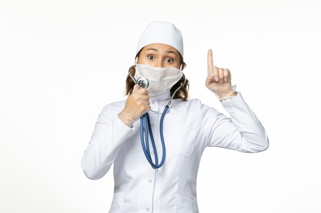 Front view young female doctor wearing protective sterile mask due to coronavirus using a stethoscope on the white surface