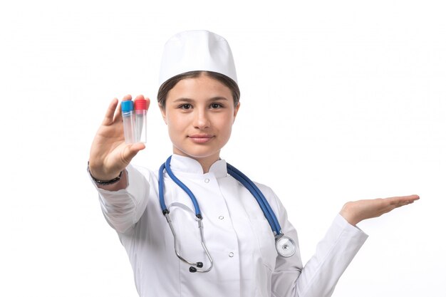 A front view young female doctor in white medical suit and white cap with blue stethoscope holding flasks 