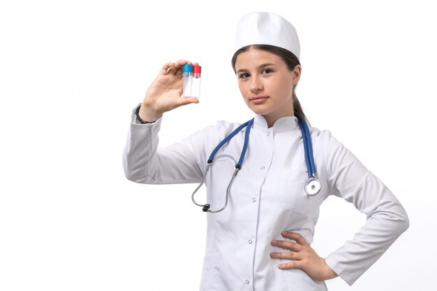 A front view young female doctor in white medical suit and white cap with blue stethoscope holding flasks 