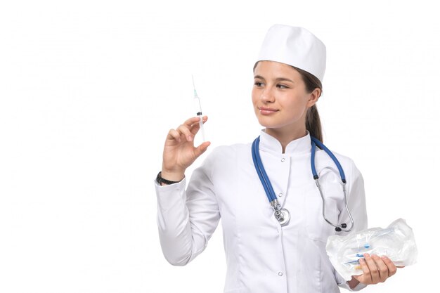 A front view young female doctor in white medical suit and white cap with blue stethoscope holding injection 