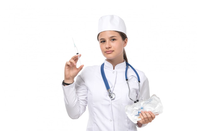 A front view young female doctor in white medical suit and white cap with blue stethoscope holding injection 