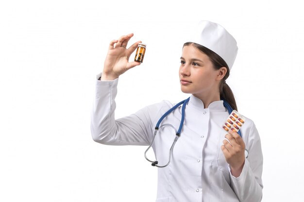 A front view young female doctor in white medical suit and white cap with blue stethoscope holding pills and potions 