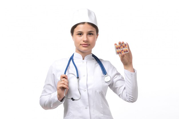 A front view young female doctor in white medical suit and white cap with blue stethoscope holding pills 