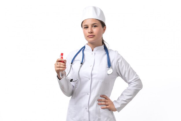 Вид спереди молодая женщина-врач в белом медицинском костюме и белой кепке с голубой стетоскоп, держа спрей