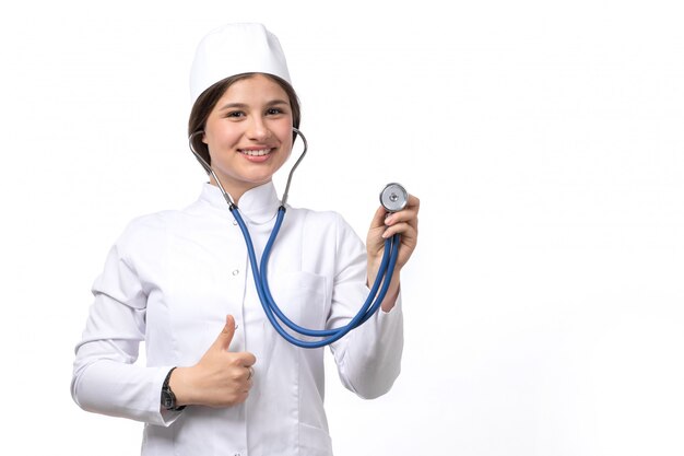 A front view young female doctor in white medical suit and white cap with blue stethoscope posing 