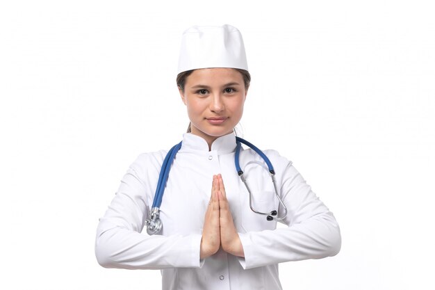 A front view young female doctor in white medical suit and white cap with blue stethoscope smiling 