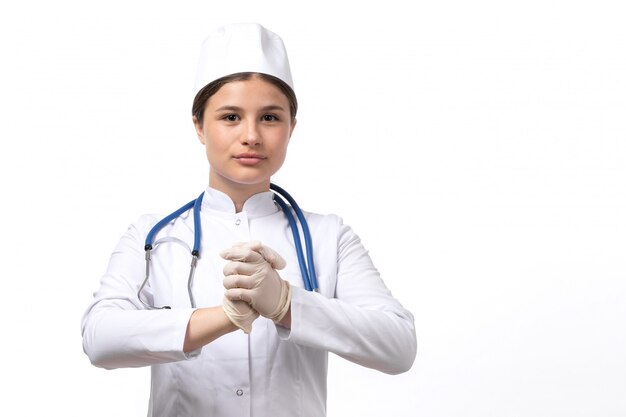 A front view young female doctor in white medical suit and white cap with blue stethoscope wearing gloves 