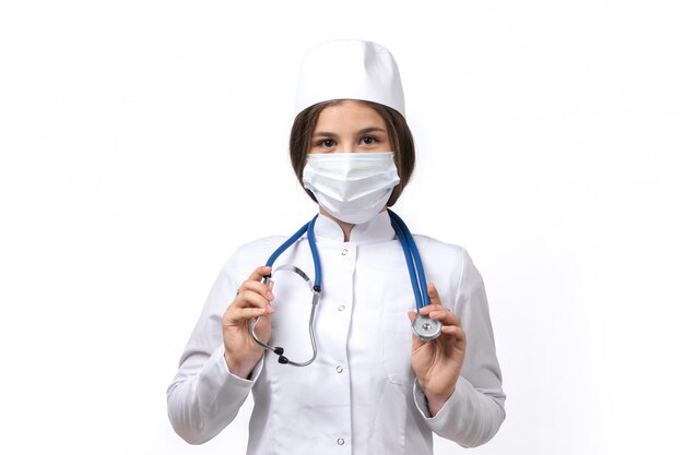 A front view young female doctor in white medical suit and white cap with blue stethoscope wearing a mask 